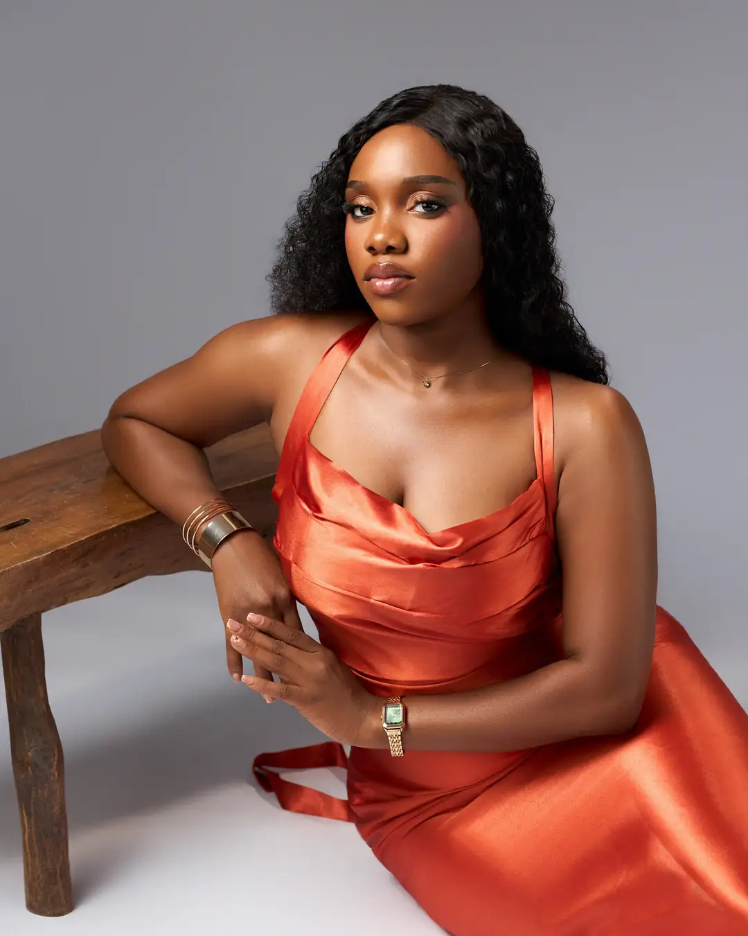 A woman in an orange satin dress poses on a wooden bench against a grey studio backdrop